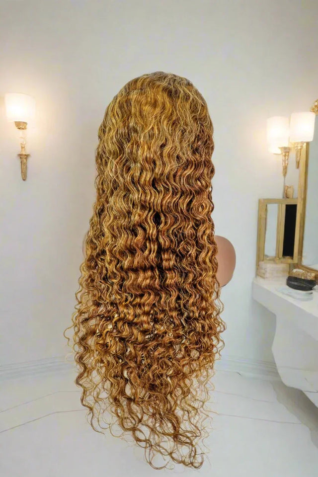 Long curly human hair wig with honey blonde highlights, displayed on mannequin in bright vanity setting