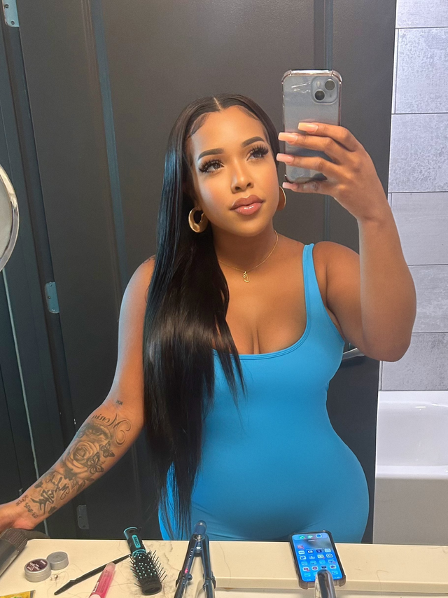 Woman taking mirror selfie with long straight human hair wig, styling tools on counter, blue bodysuit