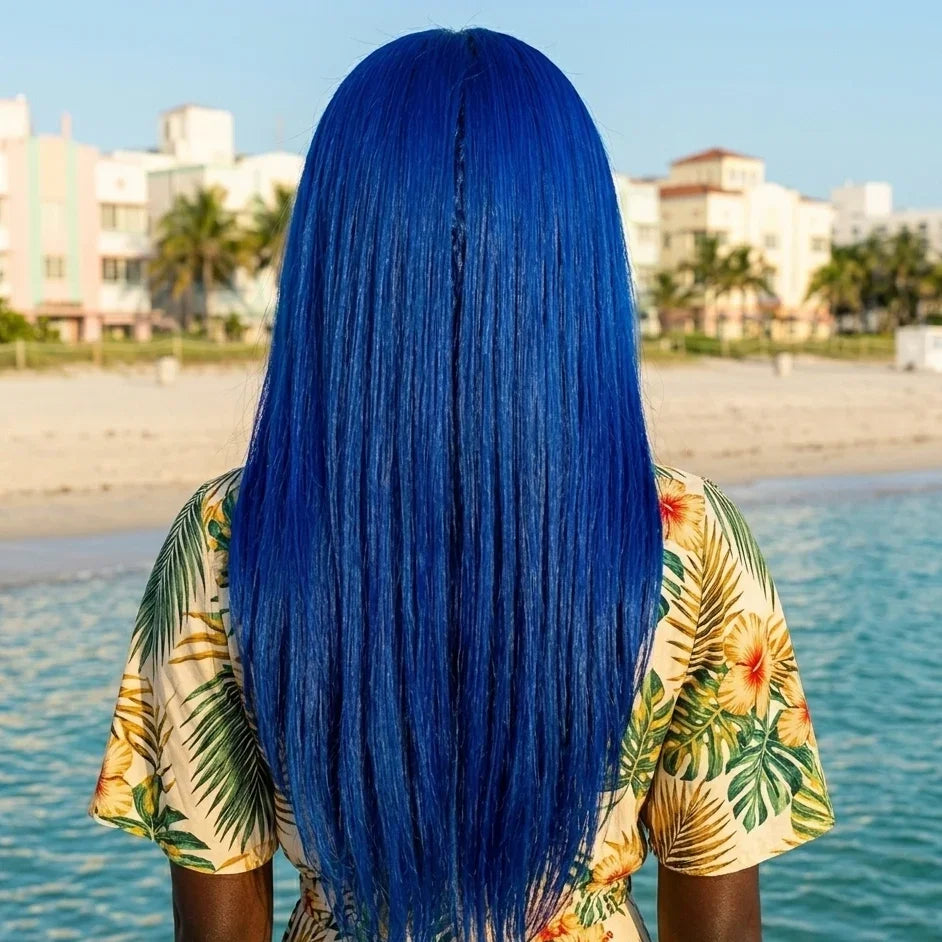 Person with blue hair standing by a beach with palm trees and buildings in the background.