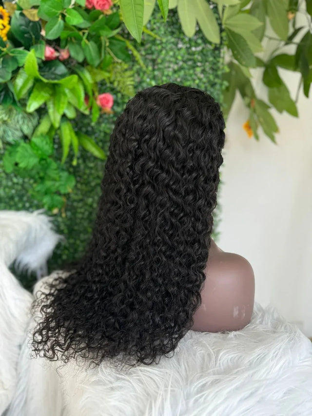 Black curly human hair wig on mannequin with lush green floral backdrop, natural styling ready