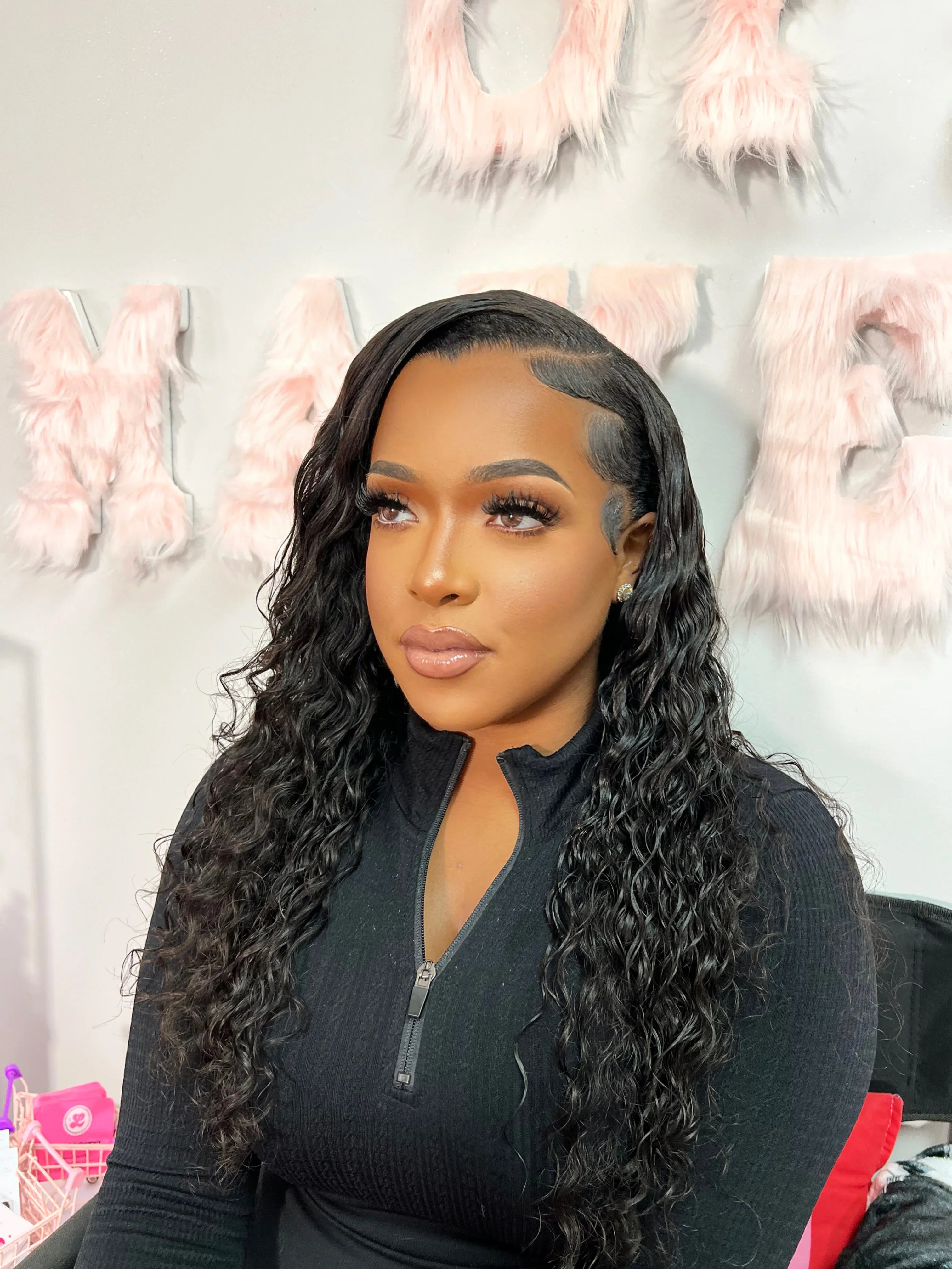 Woman wearing a natural-looking human hair wig with deep waves, styled with baby hairs, against a pink fuzzy letter backdrop.