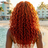Person with curly red hair standing on a beach with palm trees and buildings in the background